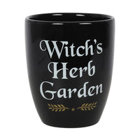 A black ceramic plant pot with the text 'WITCH'S HERB GARDEN' written in white and gold letters, along with a decorative leafy design.